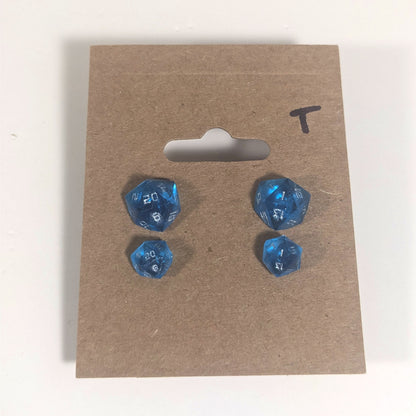 Two pairs of handmade d20 earrings with titanium hardware that are sapphire blue with silver numbers on a kraft brown earring card with a T in the top right corner. The earrings on the left of each pair centers the 20 face and the earrings on the right of each pair centers the 1 face.