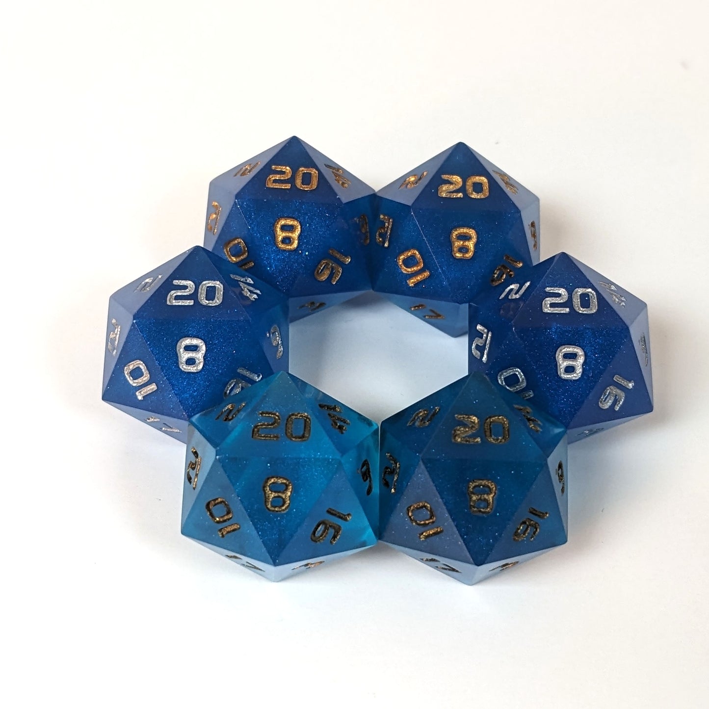 Three pairs of standard d20s are arranged in a hexagon on a white background. All of the handmade dice are a rich blue. The top two d20s have gold numbers, the middle two have silver numbers, and the bottom two have bronze numbers, showing how each number color affects the appearance of the hue of the rich blue dice.