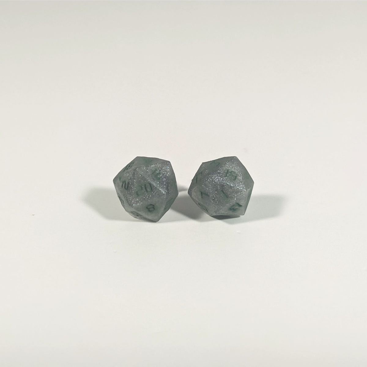 A single pair of the larger size handmade d20 earrings with titanium hardware that are dark silver with forest green numbers on a white background. The earring on the left centers the 20 face and the earring on the right centers the 1 face.