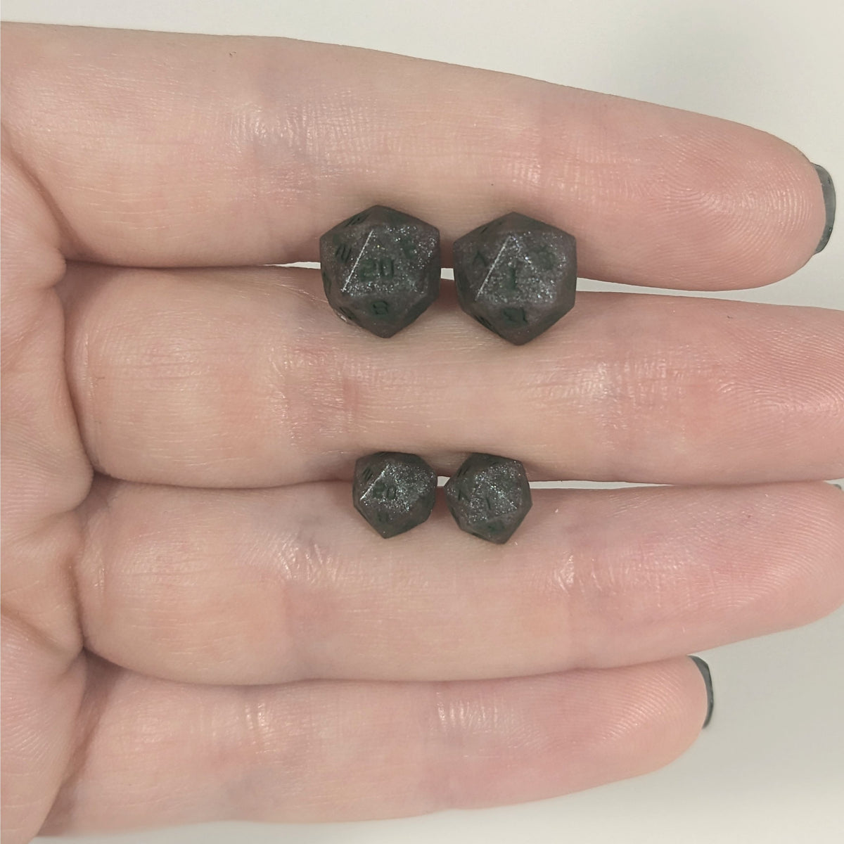 Two pairs of handmade d20 earrings with titanium hardware that are dark silver with forest green numbers held between the fingers of a flat hand. The larger pair is between the pointer and middle finger, the smaller between the middle and ring finger. The earrings on the left of each pair centers the 20 face and the earrings on the right of each pair centers the 1 face.