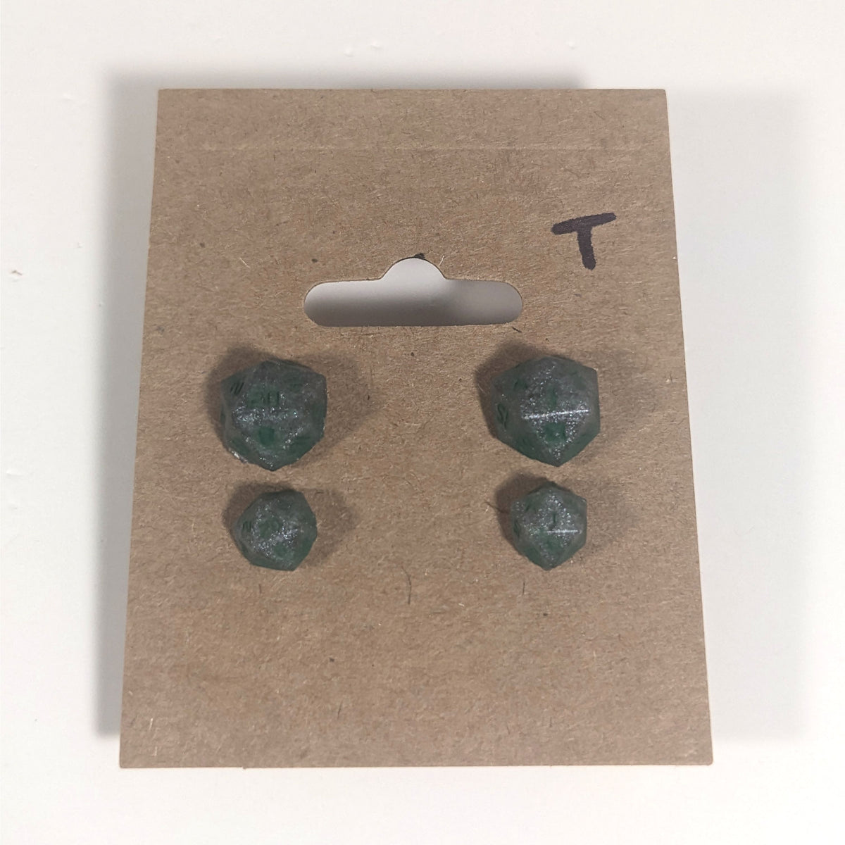 Two pairs of handmade d20 earrings with titanium hardware that are dark silver with forest green numbers on a kraft brown earring card with a T in the top right corner. The earrings on the left of each pair centers the 20 face and the earrings on the right of each pair centers the 1 face.