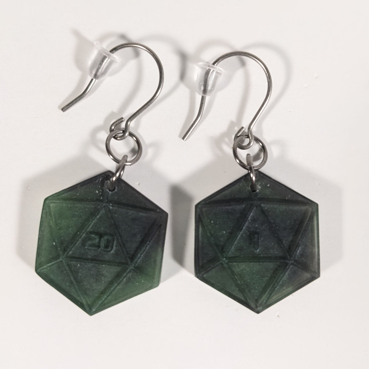 A pair of black and dark green dangly, flat d20 earrings with silver color titanium and niobium hardware and bell shaped rubber backs on a white background. The earring on the left has a 20 on it and the earring on the right has a 1 on it, both in a sci-fi style font.