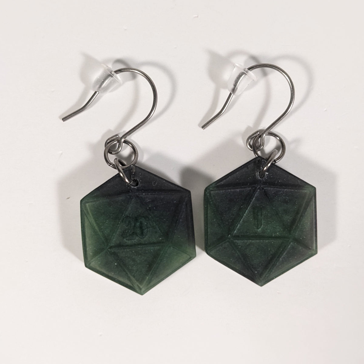 A pair of black and dark green dangly, flat d20 earrings with silver color titanium and niobium hardware and bell shaped rubber backs on a white background. The earring on the left has a 20 on it and the earring on the right has a 1 on it, both in a fantasy style font.
