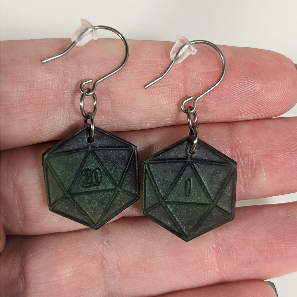 A pair of black and dark green dangly, flat d20 earrings with silver color titanium and niobium hardware with bell shaped rubber backs held on on a hand on a white background. The earring on the left has a 20 on it and the earring on the right has a 1 on it, both in a fantasy style font.