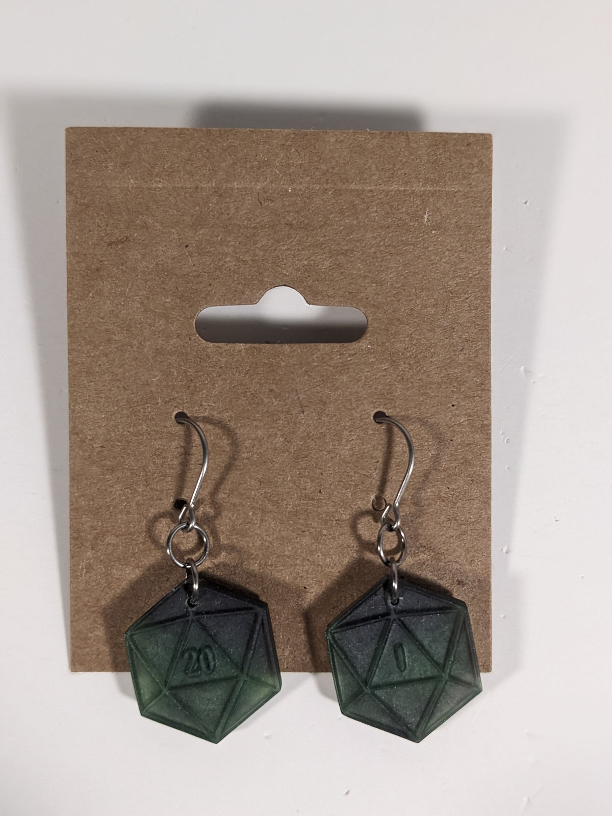 A pair of black and dark green dangly, flat d20 earrings with silver color titanium and niobium hardware on a kraft earring card on a white background. The earring on the left has a 20 on it and the earring on the right has a 1 on it, both in a fantasy style font.