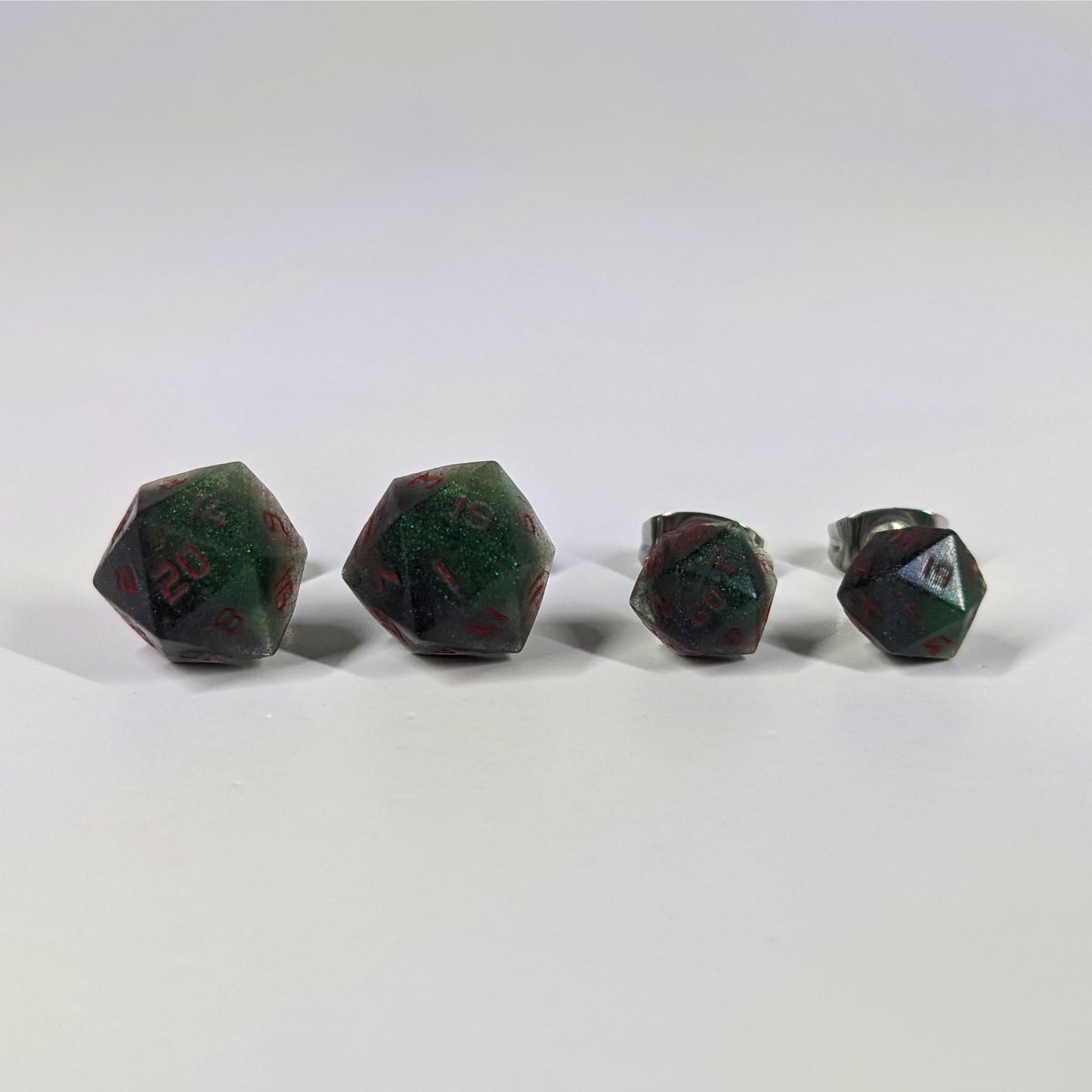 Two pairs of handmade d20 earrings that are dark green and black with red numbers on a white background sit in a line with the larger pair on the left and the smaller pair on the right. The earrings on the left of each pair centers the 20 face and the earrings on the right of each pair centers the 1 face.