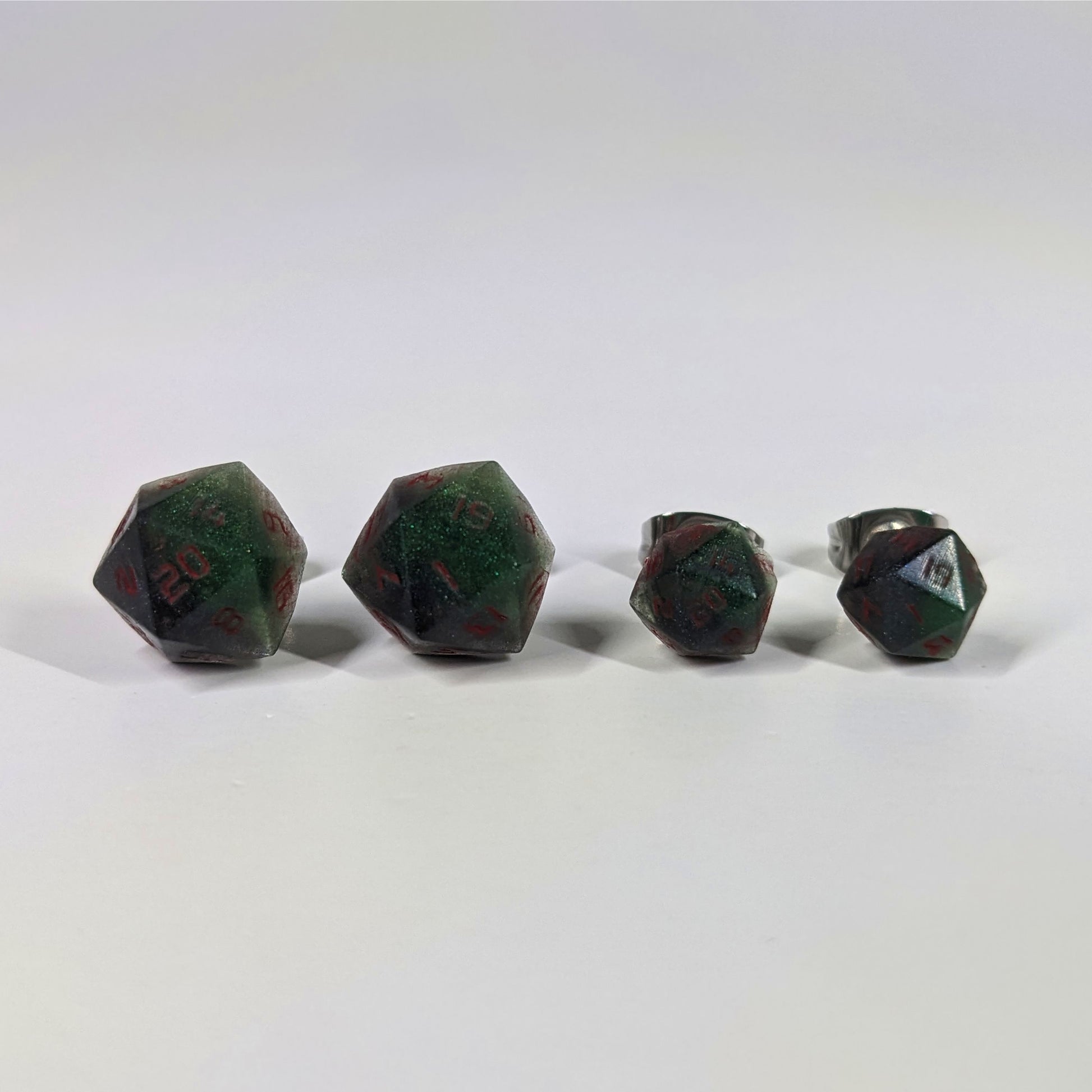 Two pairs of handmade d20 earrings that are dark green and black with red numbers on a white background sit in a line with the larger pair on the left and the smaller pair on the right. The earrings on the left of each pair centers the 20 face and the earrings on the right of each pair centers the 1 face.