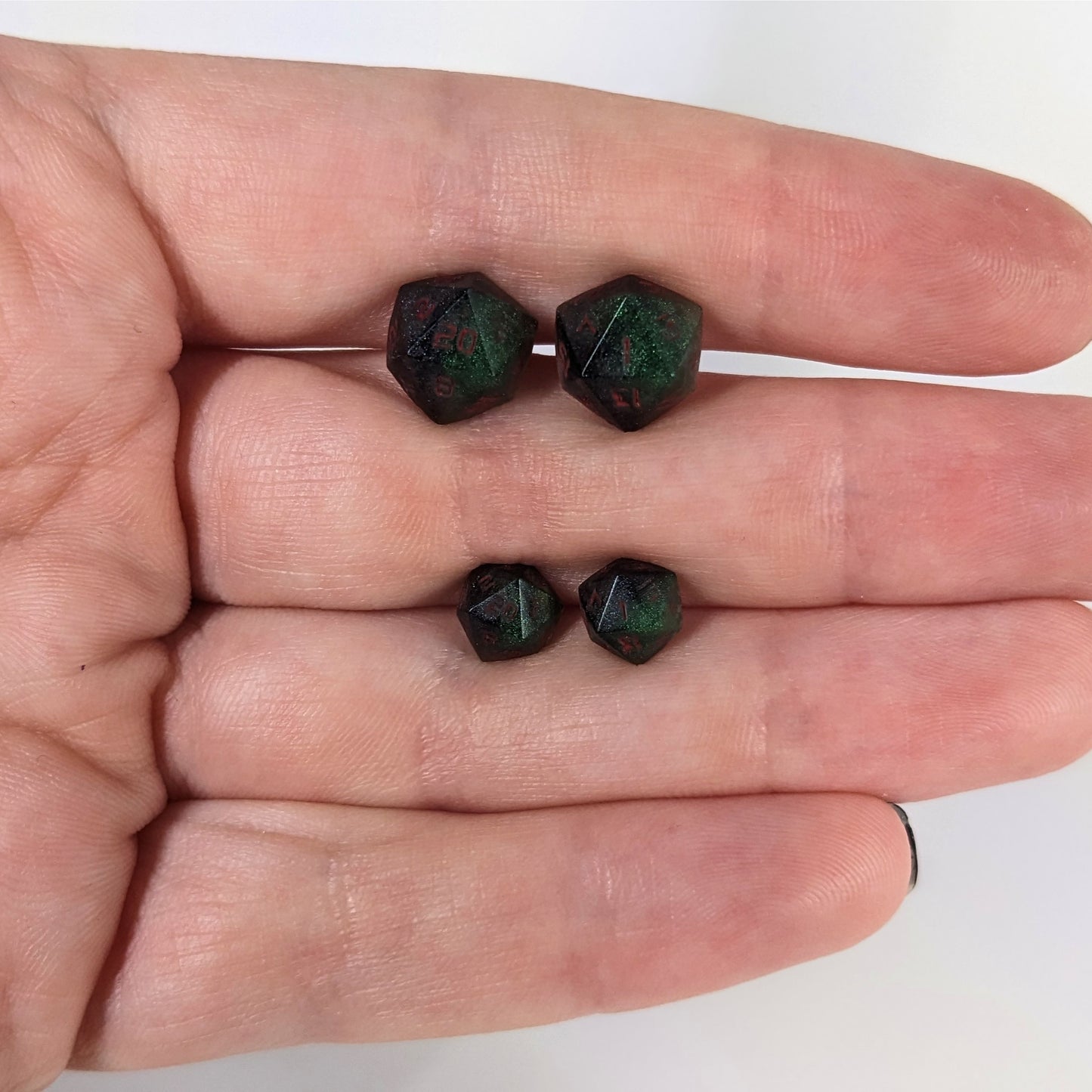 Two pairs of handmade d20 earrings that are dark green and black with red numbers held between the fingers of a flat hand. The larger pair is between the pointer and middle finger, the smaller between the middle and ring finger. The earrings on the left of each pair centers the 20 face and the earrings on the right of each pair centers the 1 face.