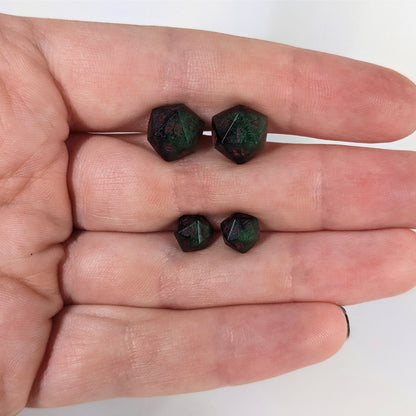 Two pairs of handmade d20 earrings that are dark green and black with red numbers held between the fingers of a flat hand. The larger pair is between the pointer and middle finger, the smaller between the middle and ring finger. The earrings on the left of each pair centers the 20 face and the earrings on the right of each pair centers the 1 face.
