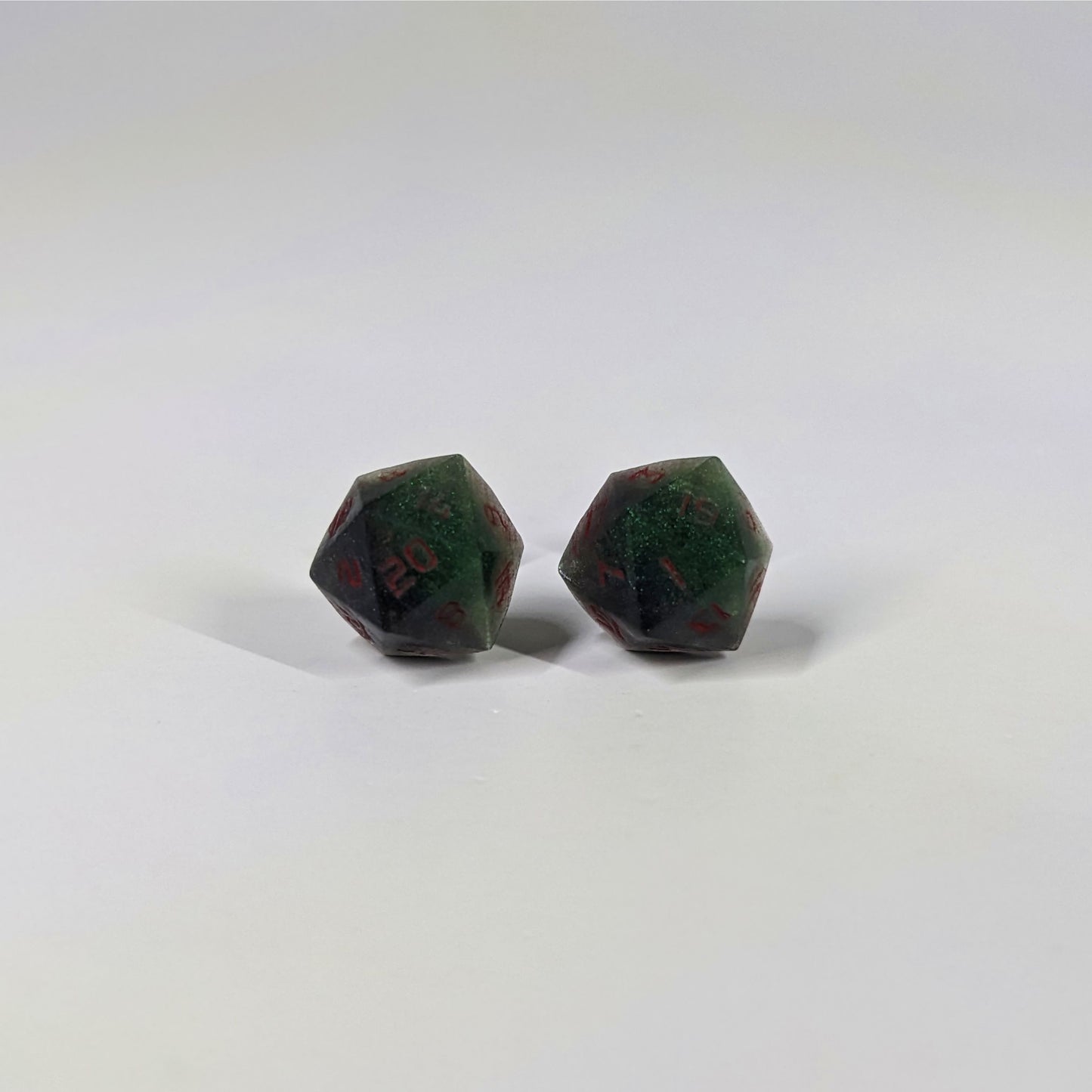 A single pair of the larger size handmade d20 earrings that are dark green and black with red numbers on a white background. The earring on the left centers the 20 face and the earring on the right centers the 1 face.