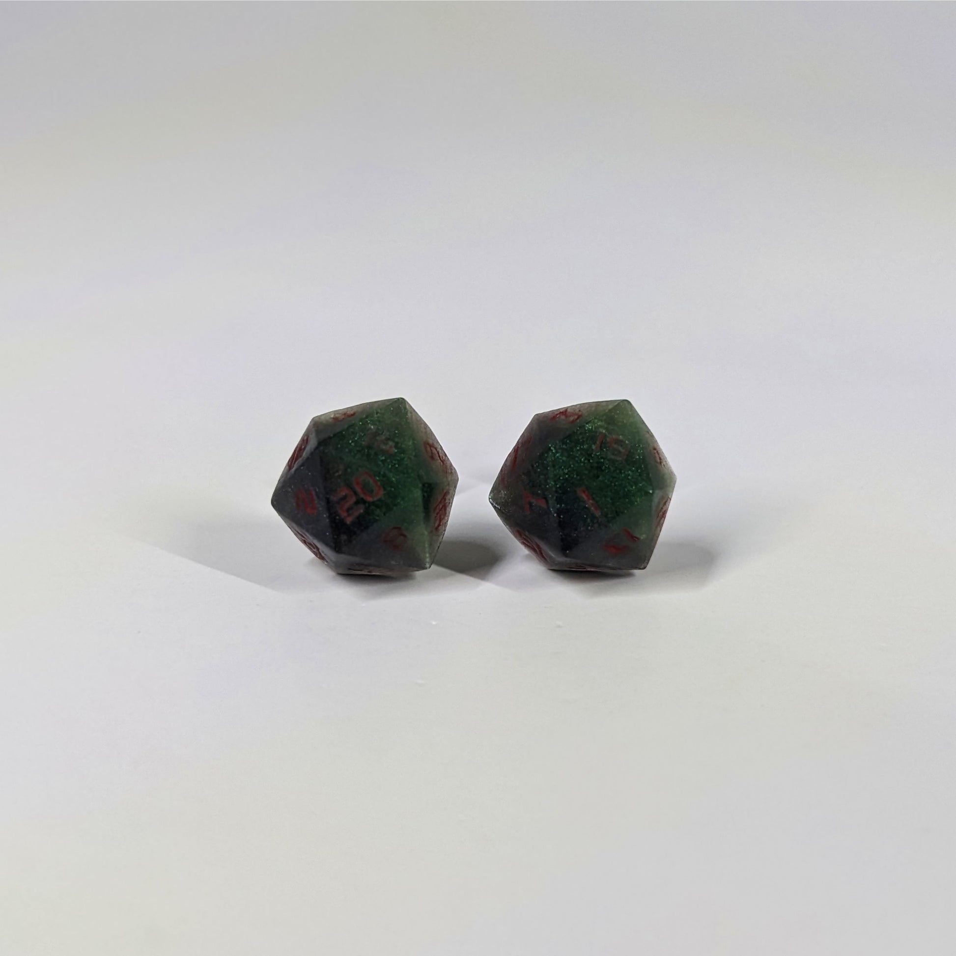 A single pair of the larger size handmade d20 earrings that are dark green and black with red numbers on a white background. The earring on the left centers the 20 face and the earring on the right centers the 1 face.