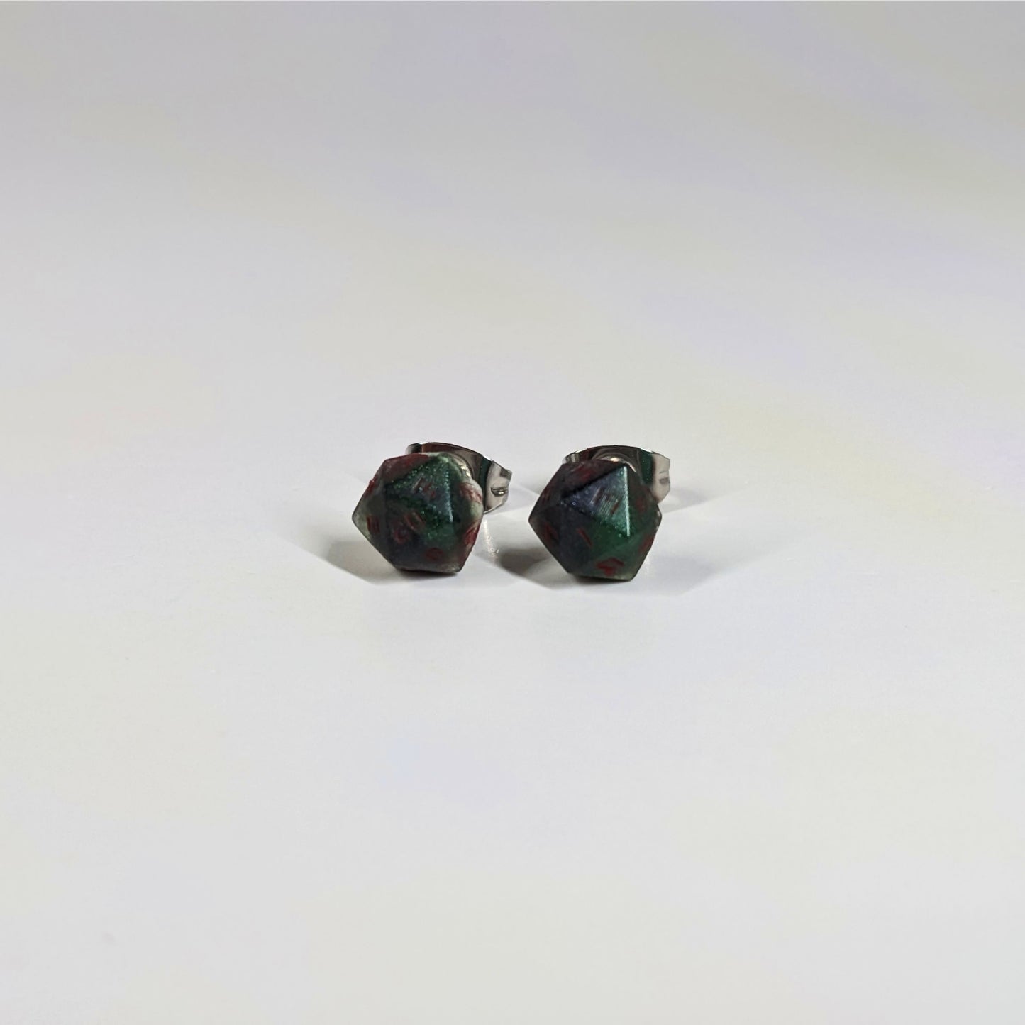 A single pair of the smaller size handmade d20 earrings that are dark green and black with red numbers on a white background. The earring on the left centers the 20 face and the earring on the right centers the 1 face.