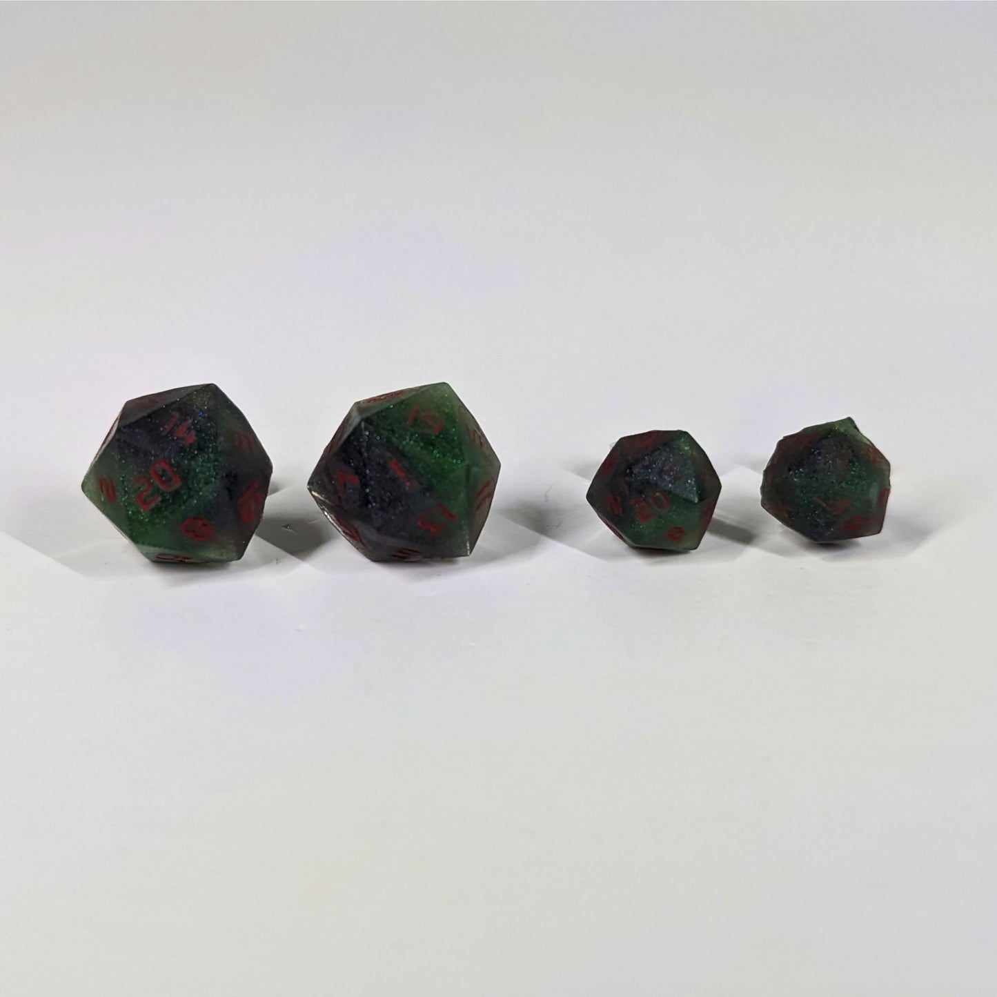 Two pairs of handmade d20 earrings with sterling silver hardware that are dark green and black with red numbers on a white background sit in a line with the larger pair on the left and the smaller pair on the right. The earrings on the left of each pair centers the 20 face and the earrings on the right of each pair centers the 1 face.