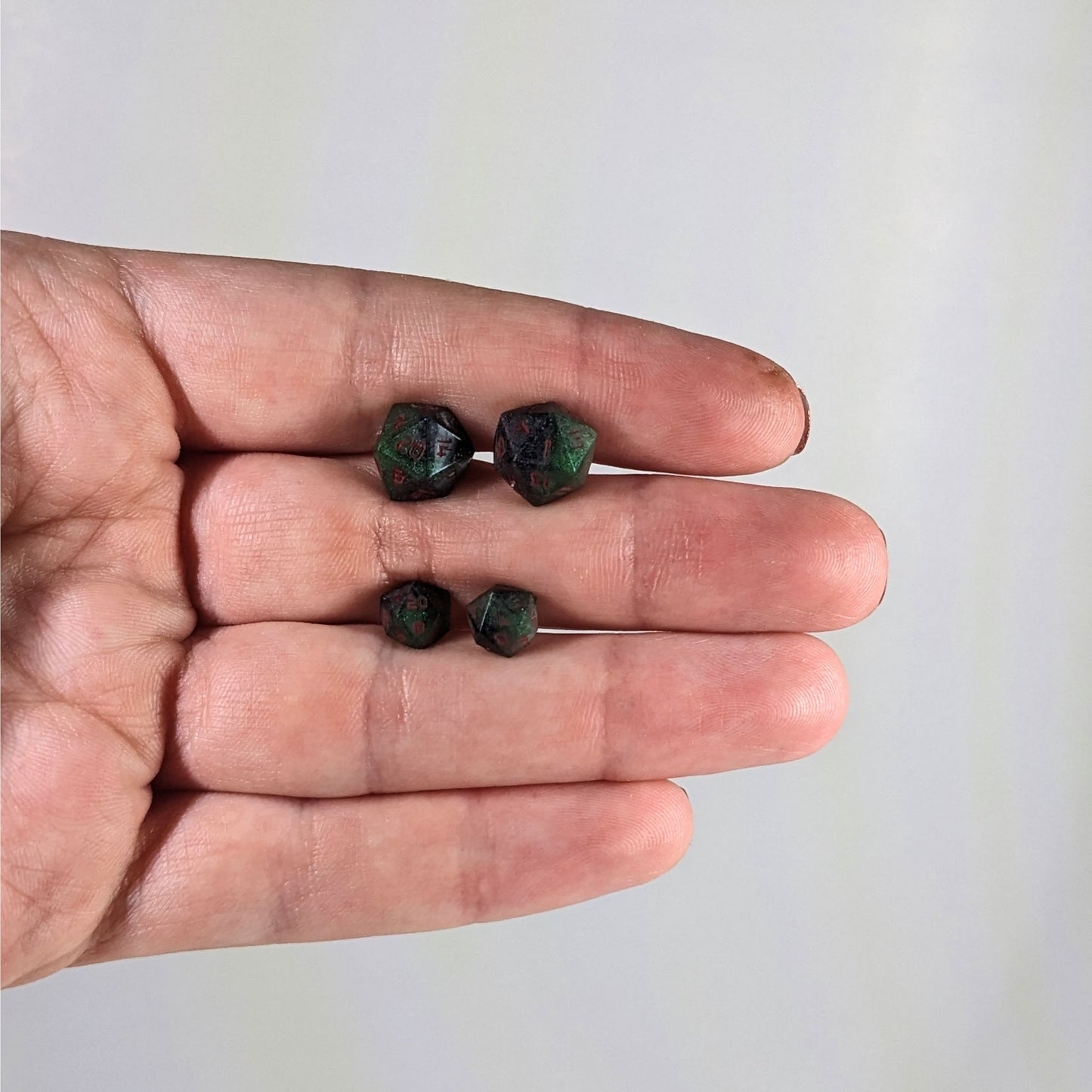 Two pairs of handmade d20 earrings with sterling silver hardware that are dark green and black with red numbers held between the fingers of a flat hand. The larger pair is between the pointer and middle finger, the smaller between the middle and ring finger. The earrings on the left of each pair centers the 20 face and the earrings on the right of each pair centers the 1 face.