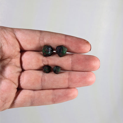 Two pairs of handmade d20 earrings with sterling silver hardware that are dark green and black with red numbers held between the fingers of a flat hand. The larger pair is between the pointer and middle finger, the smaller between the middle and ring finger. The earrings on the left of each pair centers the 20 face and the earrings on the right of each pair centers the 1 face.