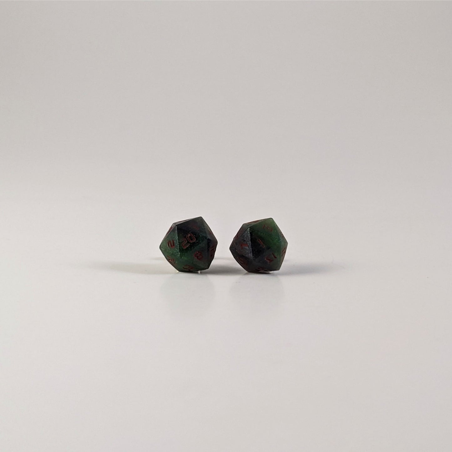 A single pair of the larger size handmade d20 earrings with sterling silver hardware that are dark green and black with red numbers on a white background. The earring on the left centers the 20 face and the earring on the right centers the 1 face.