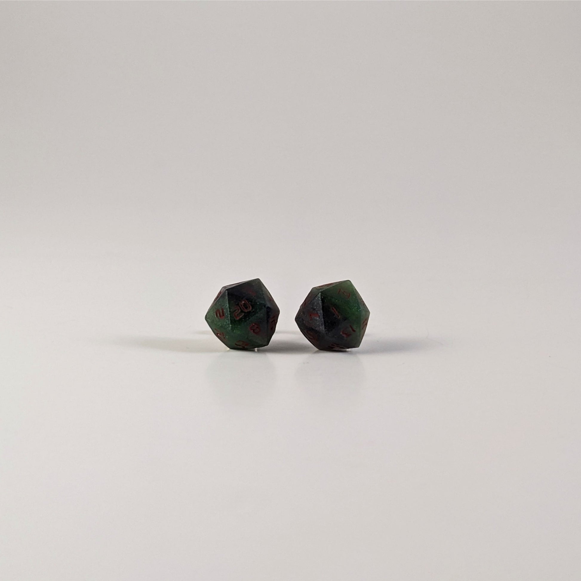 A single pair of the larger size handmade d20 earrings with sterling silver hardware that are dark green and black with red numbers on a white background. The earring on the left centers the 20 face and the earring on the right centers the 1 face.