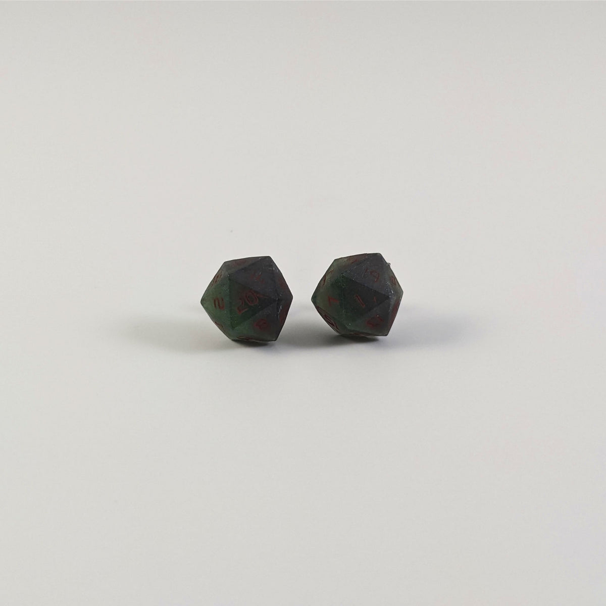 A single pair of the larger size handmade d20 earrings with titanium hardware that are dark green and black with deep red numbers on a white background. The earring on the left centers the 20 face and the earring on the right centers the 1 face.