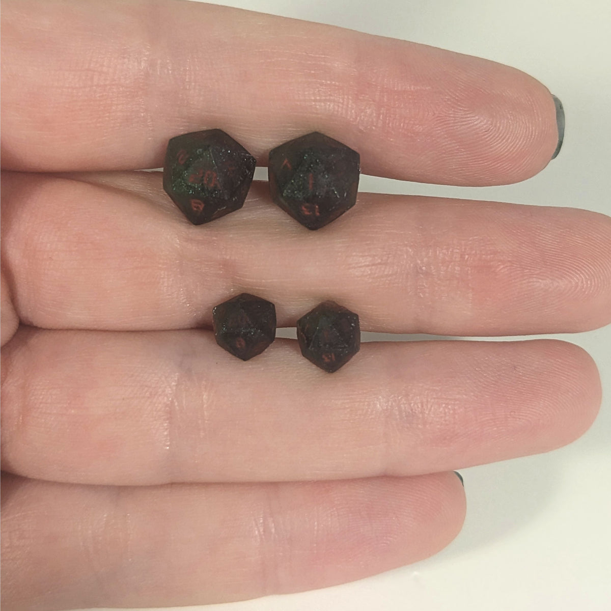 Two pairs of handmade d20 earrings with titanium hardware that are dark green and black with deep red numbers held between the fingers of a flat hand. The larger pair is between the pointer and middle finger, the smaller between the middle and ring finger. The earrings on the left of each pair centers the 20 face and the earrings on the right of each pair centers the 1 face.