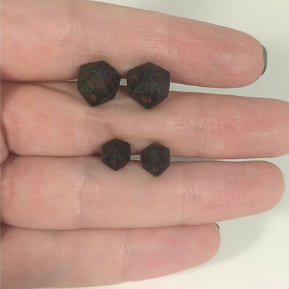 Two pairs of handmade d20 earrings with titanium hardware that are dark green and black with deep red numbers held between the fingers of a flat hand. The larger pair is between the pointer and middle finger, the smaller between the middle and ring finger. The earrings on the left of each pair centers the 20 face and the earrings on the right of each pair centers the 1 face.