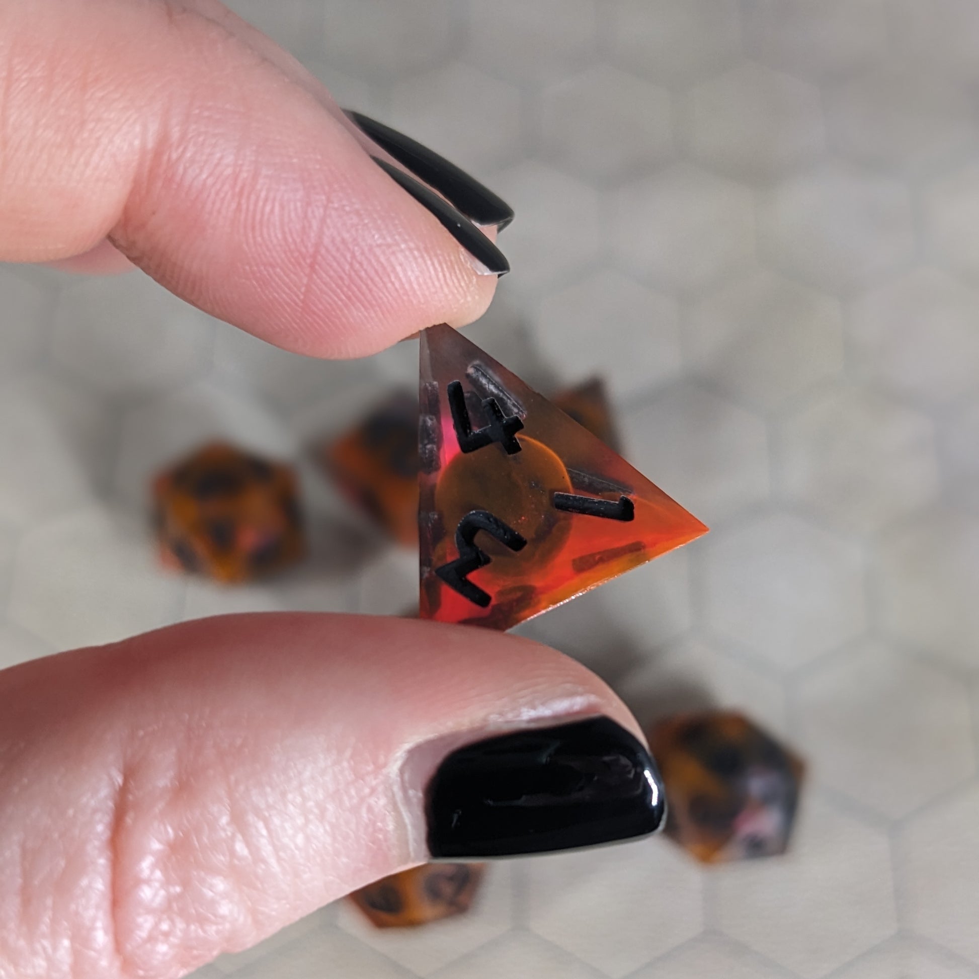 A d4 that is a glowy red-orange with a dark spherical insert and black numbers is held up to the camera to show off the red-orange swirls around the dark, spherical insert and otherwise clear shell. The face with 1, 3, and 4 in the corners is angled toward the camera.