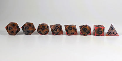 A straight on photo of a complete set of polyhedral dice that are a glowy red-orange with dark spherical inserts and black numbers arranged in a straight line. They are lined up with the 2 d20s on the left going down to the d4 on the right side. The highest value number of each die is facing forward.