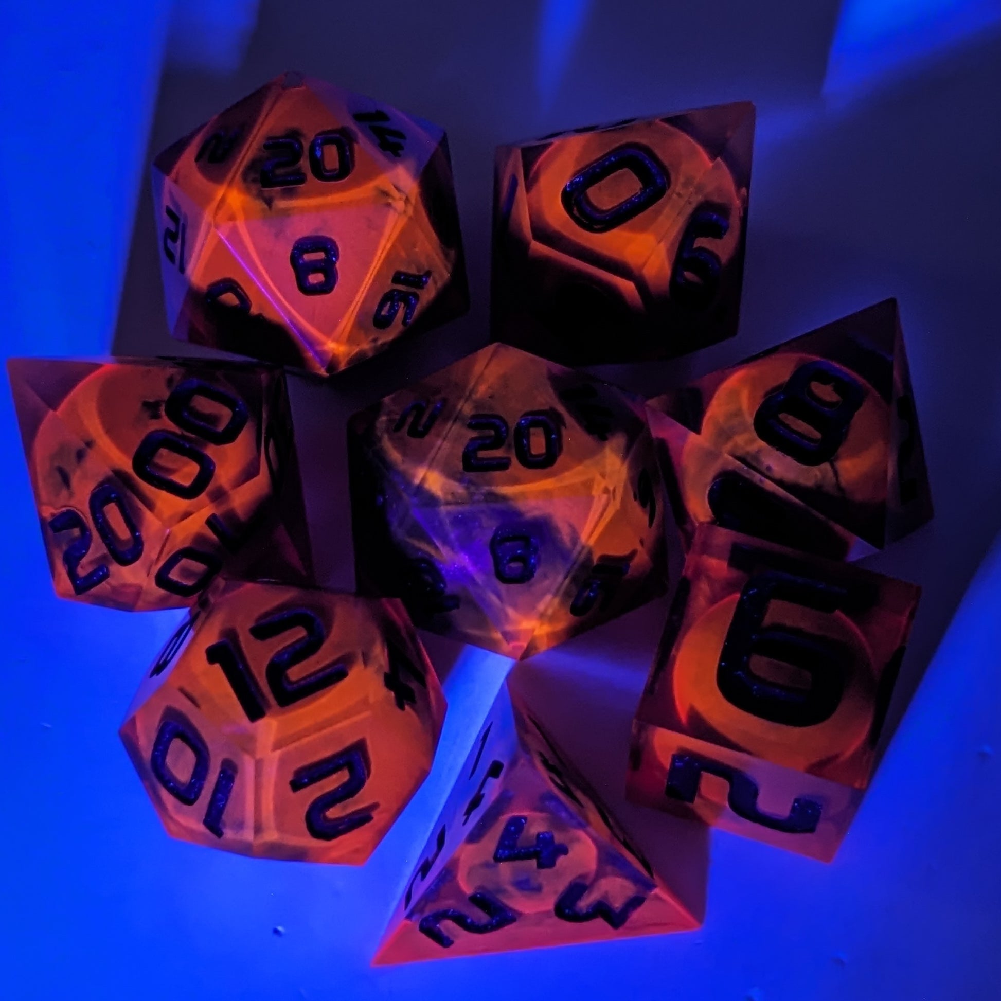 A complete set of 8 handmade artisan dice that are a glowy red-orange with dark spherical inserts and black numbers. The orange color is actually glowing as a reaction to UV light. The DnD dice set is arranged in a circle and features 2 D20s, one of which is in the center surrounded by the other 7 dice. The background is tinged blue from the UV light.