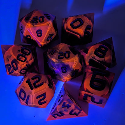 A complete set of 8 handmade artisan dice that are a glowy red-orange with dark spherical inserts and black numbers. The orange color is actually glowing as a reaction to UV light. The DnD dice set is arranged in a circle and features 2 D20s, one of which is in the center surrounded by the other 7 dice. The background is tinged blue from the UV light.