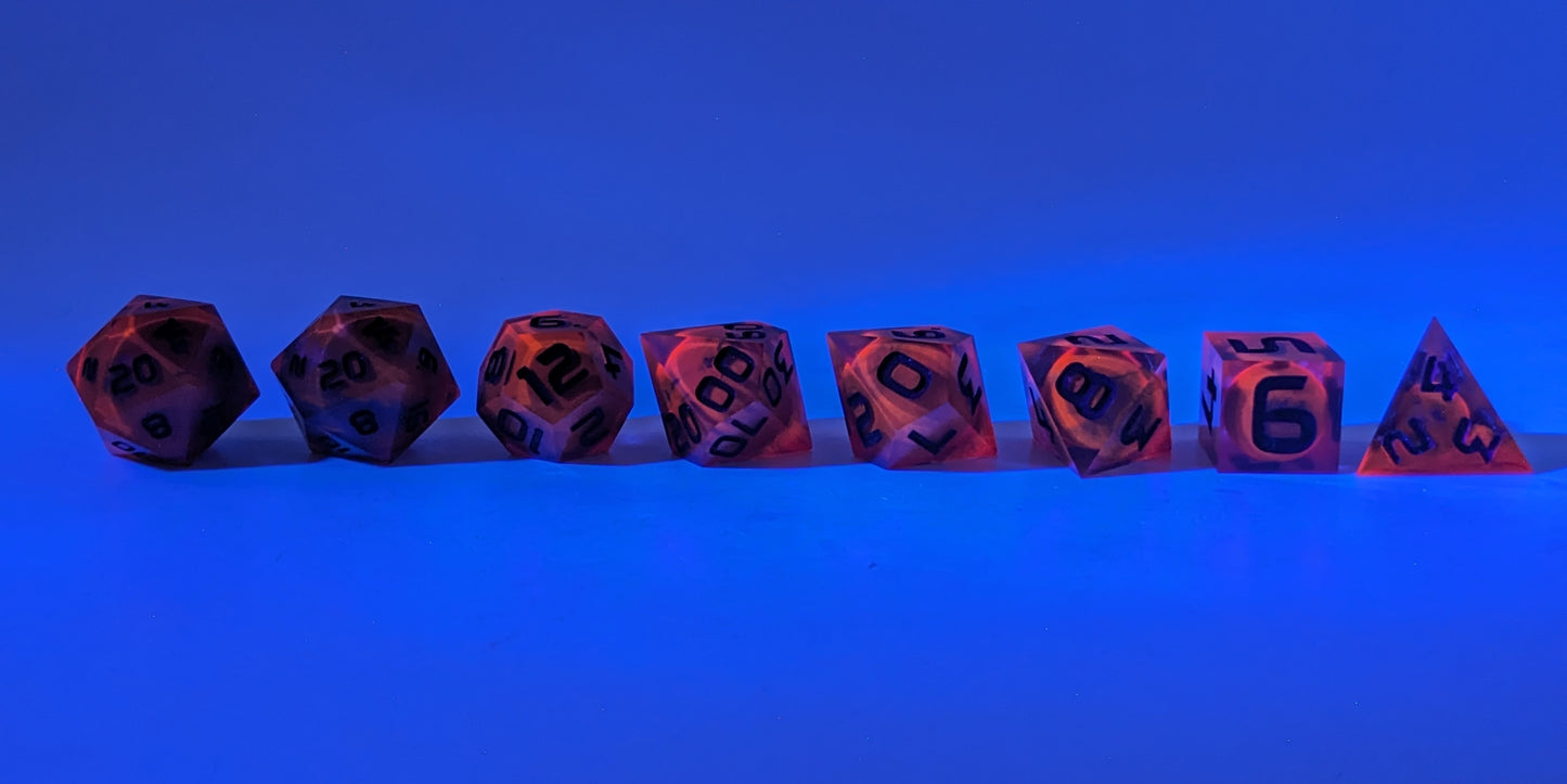 A straight on photo of a complete set of polyhedral dice that are a glowy red-orange with dark spherical inserts and black numbers arranged in a straight line. The red-orange pigment is glowing orange in reaction to a UV light. They are lined up with the 2 d20s on the left going down to the d4 on the right side. The highest value number of each die is facing forward. The background is tinged blue from the UV light.