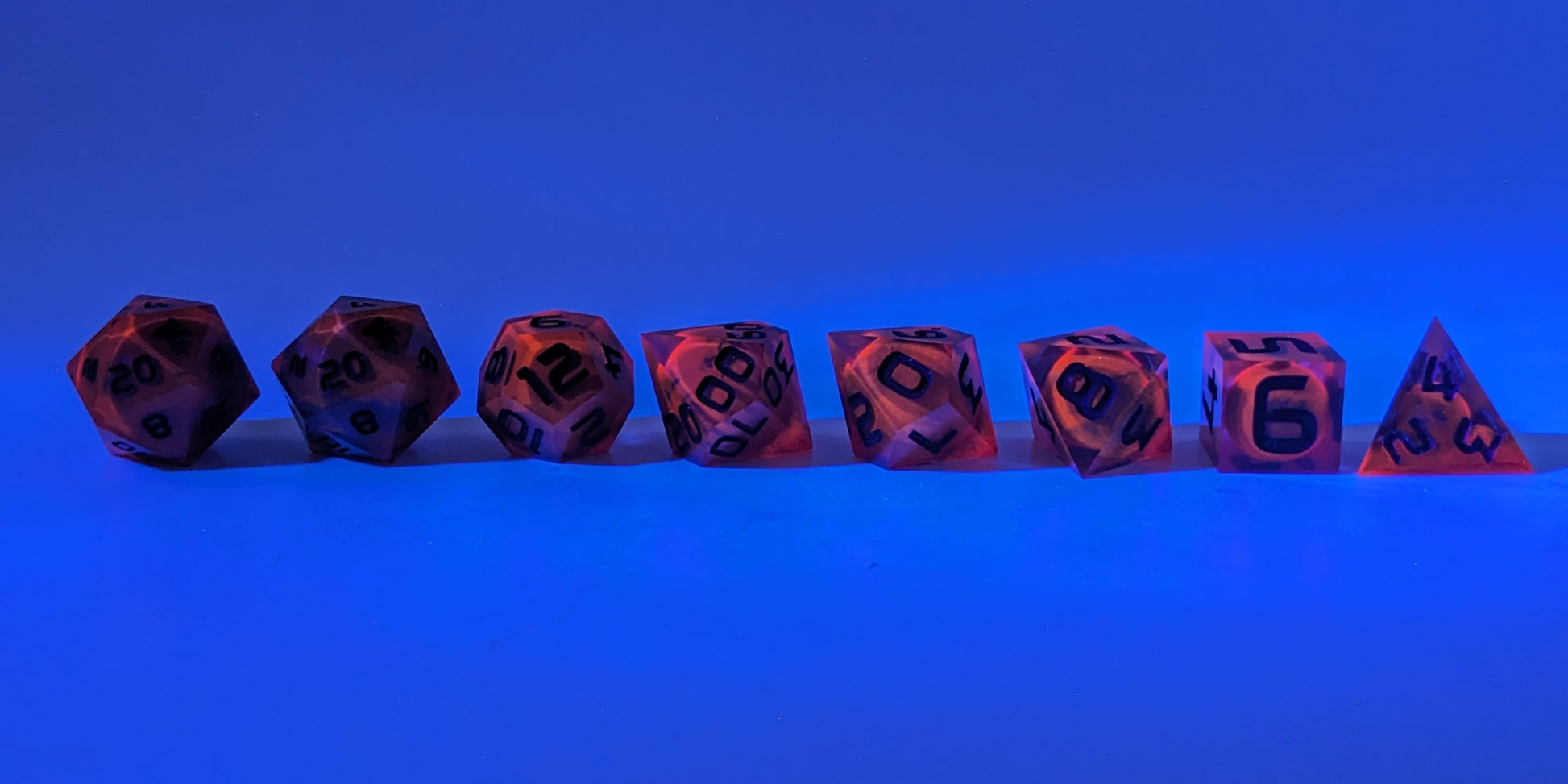 A straight on photo of a complete set of polyhedral dice that are a glowy red-orange with dark spherical inserts and black numbers arranged in a straight line. The red-orange pigment is glowing orange in reaction to a UV light. They are lined up with the 2 d20s on the left going down to the d4 on the right side. The highest value number of each die is facing forward. The background is tinged blue from the UV light.