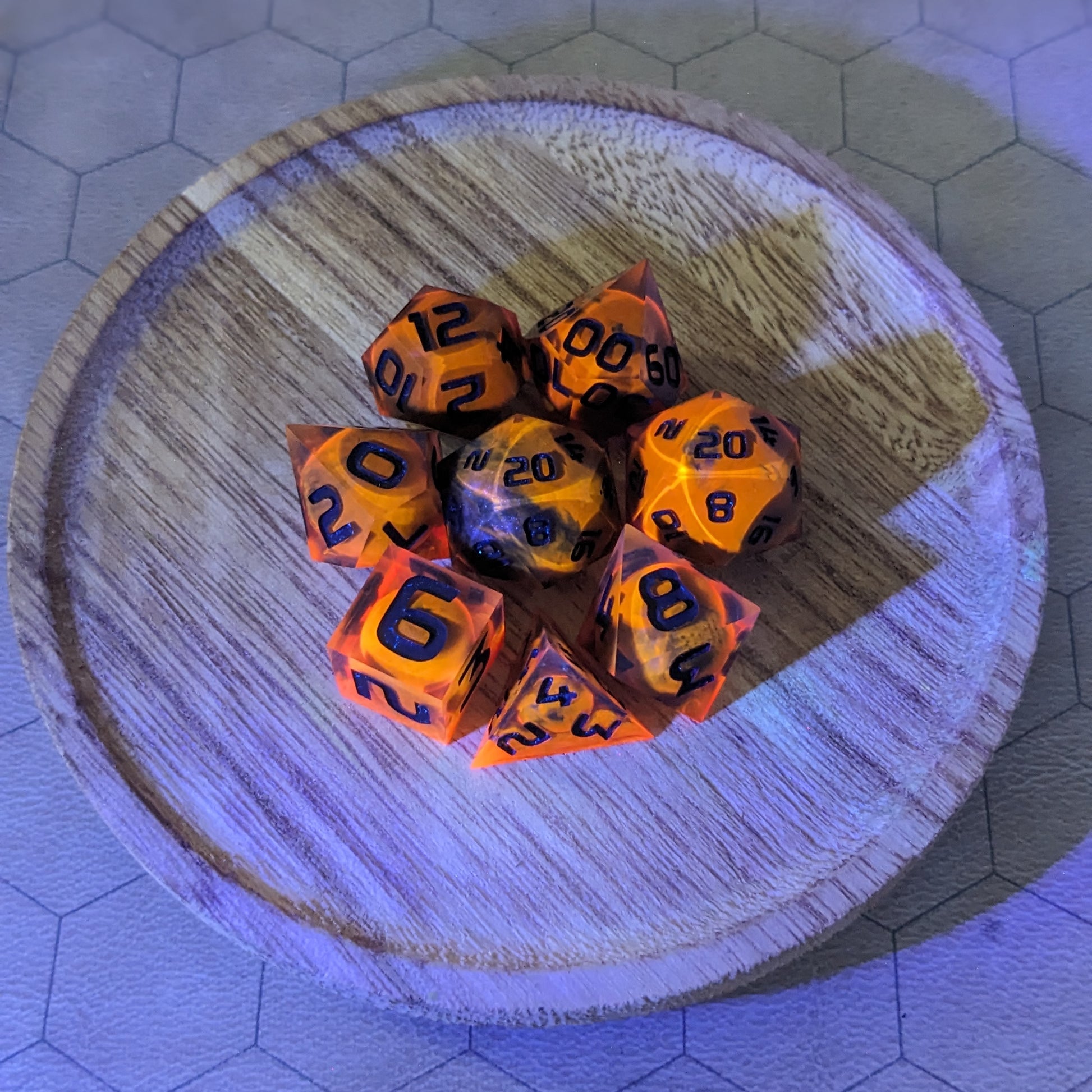A complete set of 8 handmade artisan dice that are a glowy red-orange with dark spherical inserts and black numbers. The orange color is actually glowing as a reaction to UV light. The DnD dice set is on a wooded circle and the dice are arranged in a circle and features 2 D20s, one of which is in the center surrounded by the other 7 dice. The background is tinged blue from the UV light.