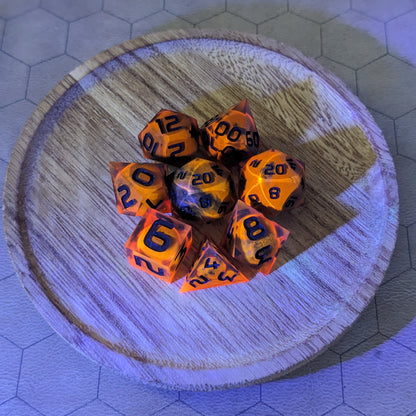 A complete set of 8 handmade artisan dice that are a glowy red-orange with dark spherical inserts and black numbers. The orange color is actually glowing as a reaction to UV light. The DnD dice set is on a wooded circle and the dice are arranged in a circle and features 2 D20s, one of which is in the center surrounded by the other 7 dice. The background is tinged blue from the UV light.