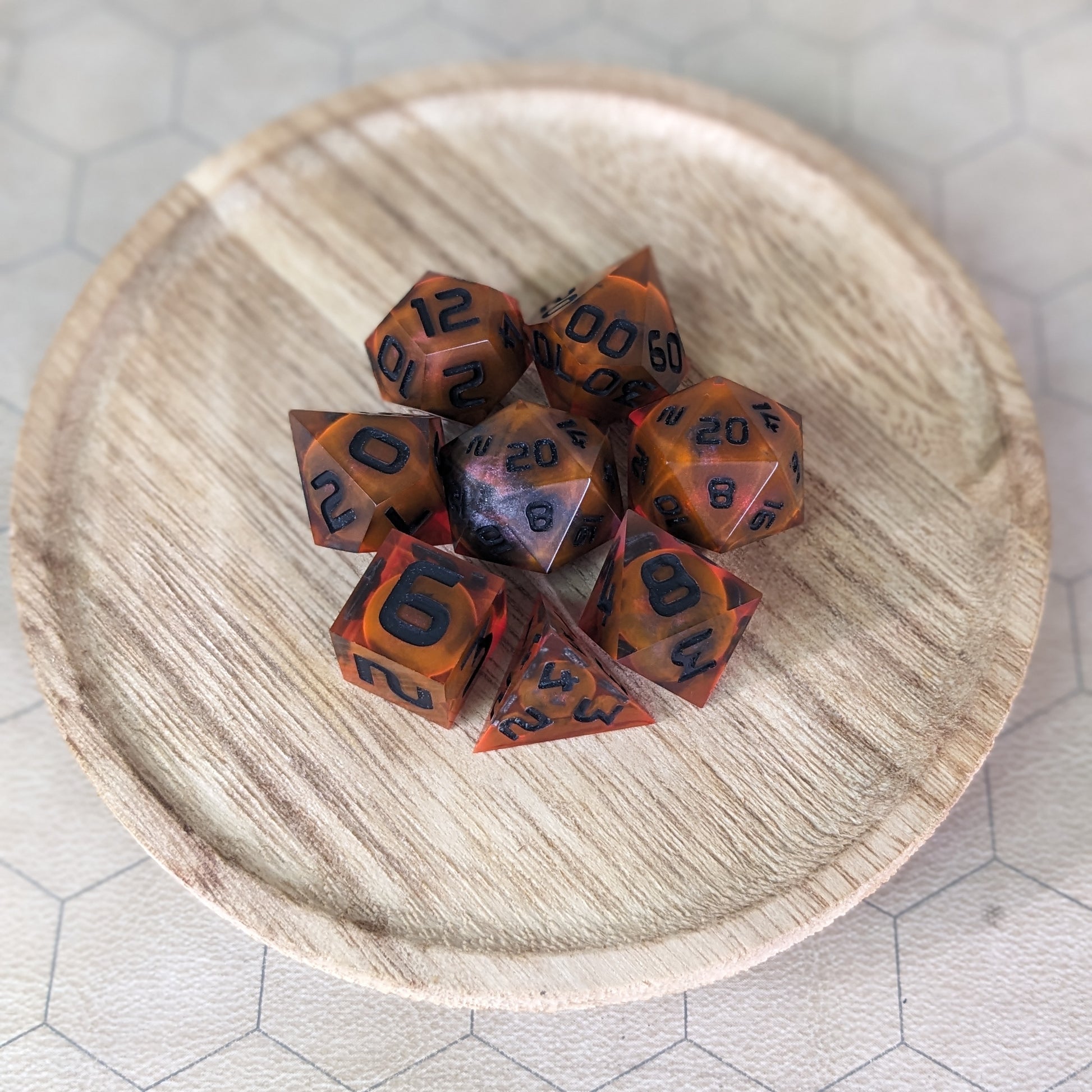  A complete set of 8 handmade artisan dice that are a glowy red-orange with dark spherical inserts and black numbers. The DnD dice set is on a wooded circle and the dice are arranged in a circle and features 2 D20s, one of which is in the center surrounded by the other 7 dice.