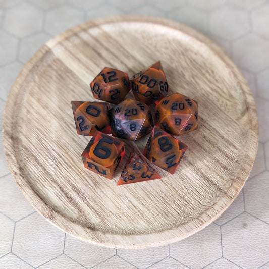  A complete set of 8 handmade artisan dice that are a glowy red-orange with dark spherical inserts and black numbers. The DnD dice set is on a wooded circle and the dice are arranged in a circle and features 2 D20s, one of which is in the center surrounded by the other 7 dice.
