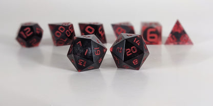 A straight on photo of a standard set of DnD dice that have dark, spherical inserts with swirls of red in the shell and red numbers focusing on 2 d20s in the center. The d20 on the left has the 1 face centered and the d20 on the right has the 20 face centered. The other 6 dice are lined up in the background but are out of focus, while the d20s are in sharp focus.