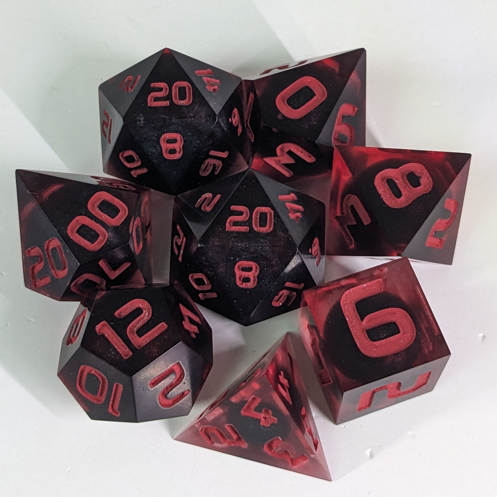 A complete set of 8 handmade artisan dice that have dark, spherical inserts with swirls of red in the shell and red numbers. The DnD dice set is arranged in a circle and features 2 D20s, one of which is in the center surrounded by the other 7 dice.