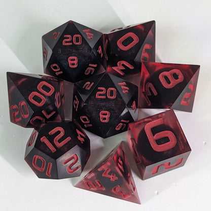 A complete set of 8 handmade artisan dice that have dark, spherical inserts with swirls of red in the shell and red numbers. The DnD dice set is arranged in a circle and features 2 D20s, one of which is in the center surrounded by the other 7 dice.
