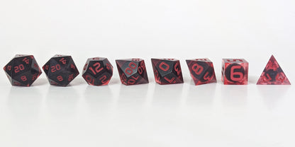 A straight on photo of a complete set of polyhedral dice that have dark, spherical inserts with swirls of red in the shell and red numbers arranged in a straight line. They are lined up with the 2 d20s on the left going down to the d4 on the right side. The highest value number of each die is facing forward.