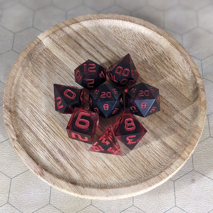 A complete set of 8 handmade artisan dice that have dark, spherical inserts with swirls of red in the shell and red numbers. The DnD dice set is on a wooded circle and the dice are arranged in a circle and features 2 D20s, one of which is in the center surrounded by the other 7 dice.