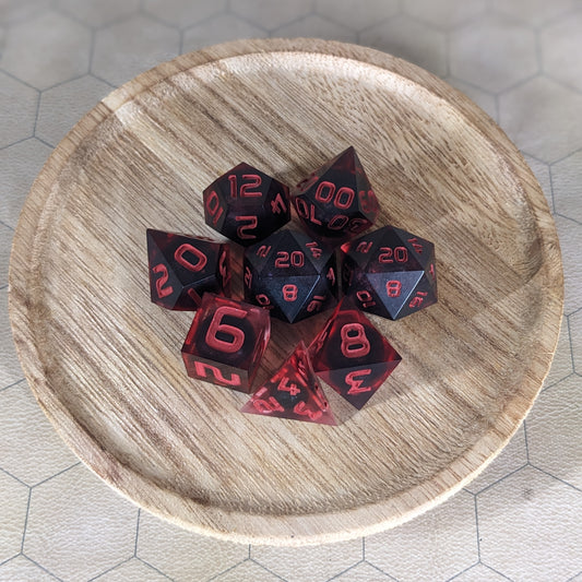 A complete set of 8 handmade artisan dice that have dark, spherical inserts with swirls of red in the shell and red numbers. The DnD dice set is on a wooded circle and the dice are arranged in a circle and features 2 D20s, one of which is in the center surrounded by the other 7 dice.
