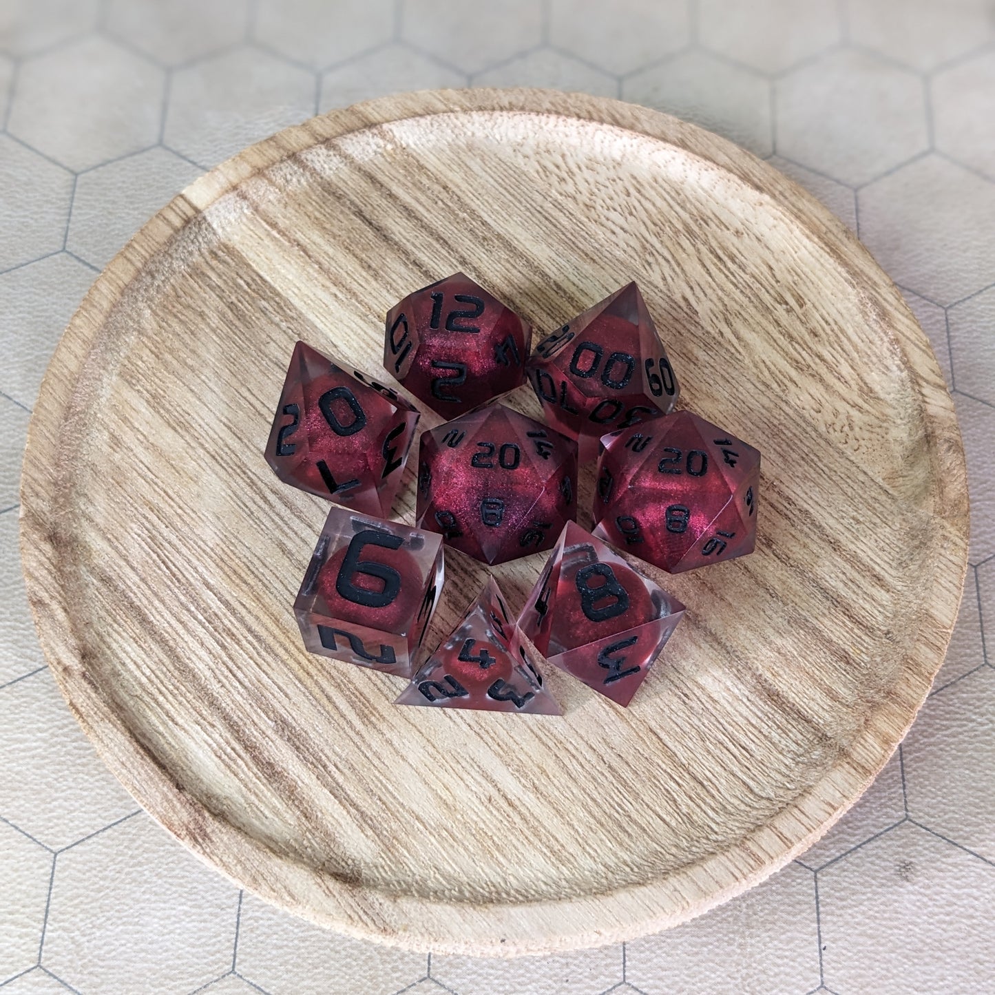 A complete set of 8 handmade artisan dice that are clear with red, spherical inserts and black numbers. The DnD dice set is on a wooded circle and the dice are arranged in a circle and features 2 D20s, one of which is in the center surrounded by the other 7 dice.