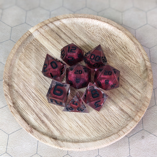 A complete set of 8 handmade artisan dice that are clear with red, spherical inserts and black numbers. The DnD dice set is on a wooded circle and the dice are arranged in a circle and features 2 D20s, one of which is in the center surrounded by the other 7 dice.