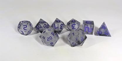 A straight on photo of a standard set of DnD dice that have black swirls around a cloudy, spherical insert and purple numbers focusing on 2 d20s in the center. The d20 on the left has the 20 face centered and the d20 on the right has the 1 face centered. The other 6 dice are lined up in the background.