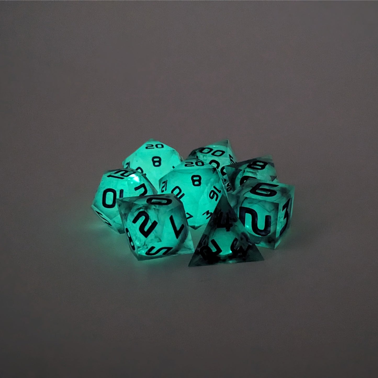 A complete set of 8 polyhedral dice that with spherical inserts glowing green, black swirls and purple numbers, showing the glow-in-the-dark feature of this dice set. The DnD dice set is arranged in a circle and features 2 D20s, one of which is in the center surrounded by the other 7 dice.