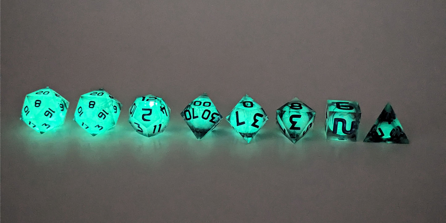 A straight on photo of a complete set of polyhedral dice that have glowing green spherical inserts and have purple numbers arranged in a straight line. They are lined up with the 2 d20s on the left going down to the d4 on the right side. The highest value number of each die is facing up. This image shows the glow-in-the-dark feature of this dice set and that you can still see the black swirls through the glow.