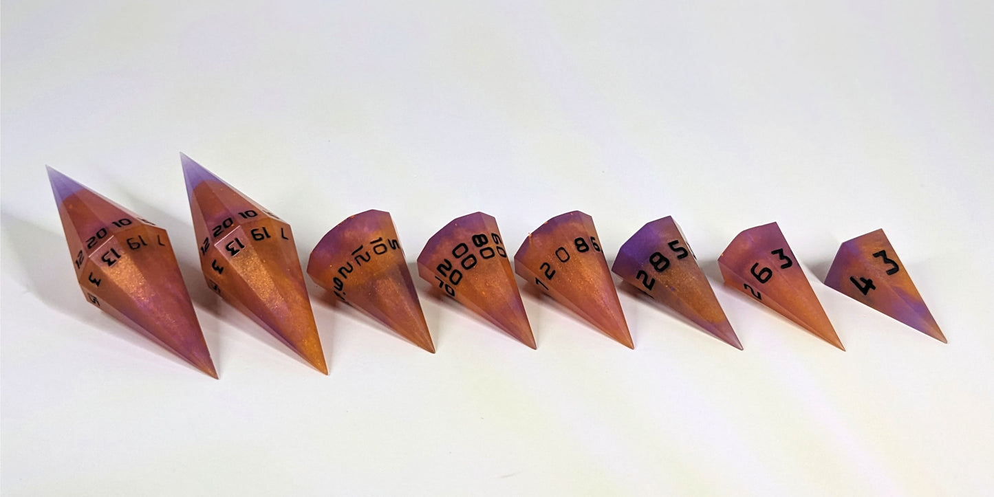 A straight on photo of a complete set of dagger DnD dice that are purple and orange with black numbers arranged in a straight line with points angled toward the right and slightly forward. They are lined up with the 2 d20s on the left going down to the d4 on the right side. The highest value number of each die is facing forward.