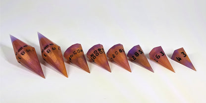 A straight on photo of a complete set of dagger DnD dice that are purple and orange with black numbers arranged in a straight line with points angled toward the right and slightly forward. They are lined up with the 2 d20s on the left going down to the d4 on the right side. The highest value number of each die is facing forward.