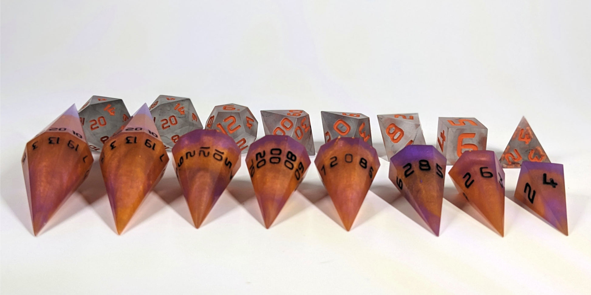 A straight on photo of a complete set of dagger dice that are purple and orange with black numbers arranged in a straight line with points angled forward. They are lined up with the 2 d20s on the left going down to the d4 on the right side. Behind the dagger set is a standard set of polyhedral dnd dice to allow for size comparison. The dagger dice are much larger overall, but not taller. The highest value number of each die is facing up.