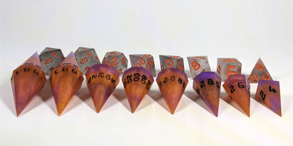 A straight on photo of a complete set of dagger dice that are purple and orange with black numbers arranged in a straight line with points angled forward. They are lined up with the 2 d20s on the left going down to the d4 on the right side. Behind the dagger set is a standard set of polyhedral dnd dice to allow for size comparison. The dagger dice are much larger overall, but not taller. The highest value number of each die is facing up.