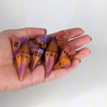 An 8 dice set of dagger style dice that are purple and orange with black numbers are held in a hand. The image shows that the set just fits in a hand, making them overall larger than a standard set of dnd dice.